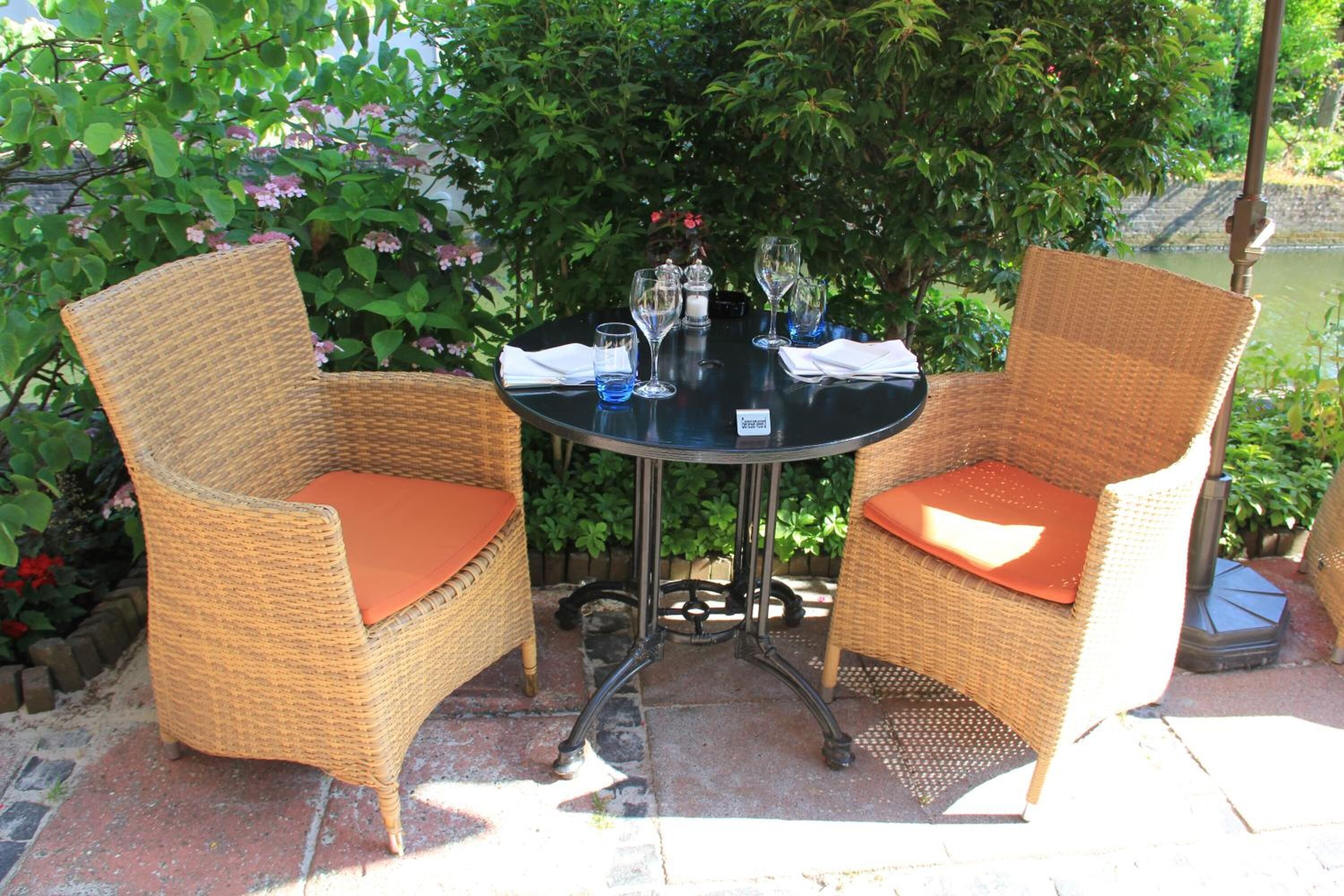 Een ronde tafel met twee rieten stoelen, elk met een oranje kussen. De tafel staat omringd door begroeiing en water.