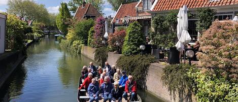 Un canal étroit où navigue un bateau transportant des passagers, le tout bordé de bâtiments traditionnels aux toits de tuiles rouges et d’une végétation luxuriante.