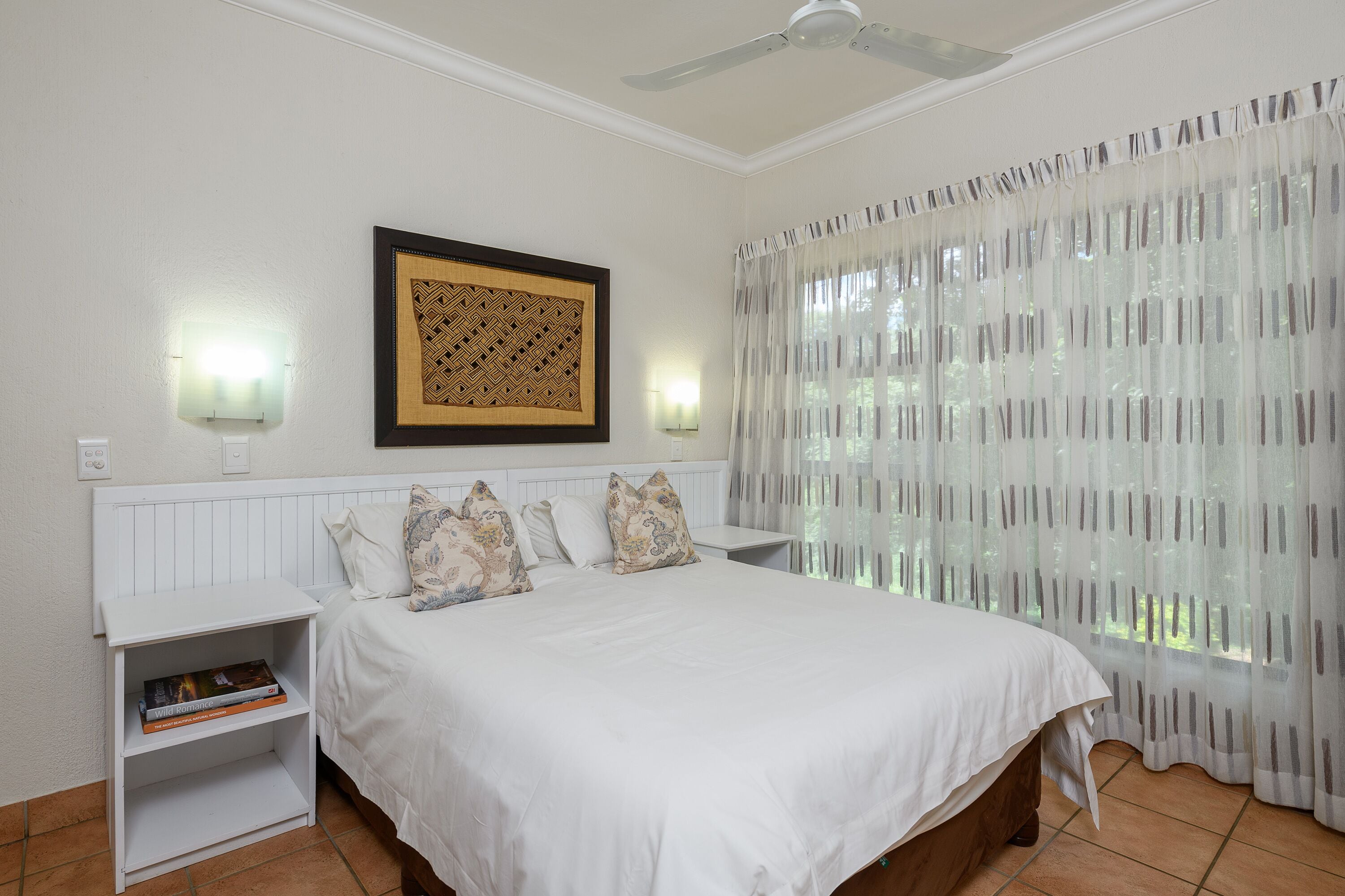 Un dormitorio con cama, mesita de noche, lámpara de pared, ventilador de techo y cortinas traslúcidas.