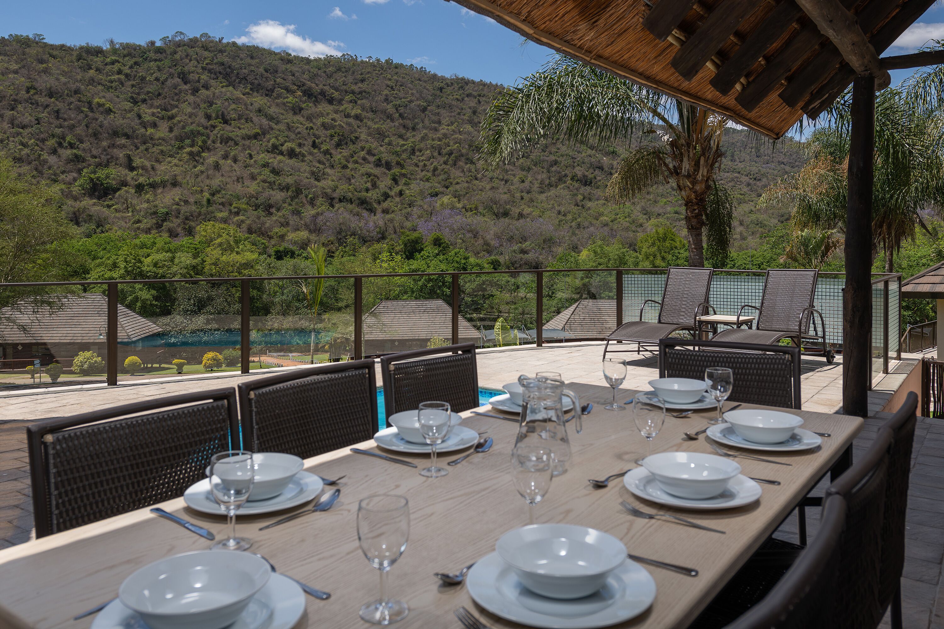 A dining area with a table set for six, surrounded by chairs, overlooking a lush landscape with a pool and a mountain in the background.