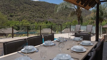 A dining area with a table set for six, surrounded by chairs, overlooking a lush landscape with a pool and a mountain in the background.