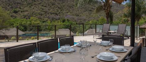 A dining area with a table set for six, surrounded by chairs, overlooking a lush landscape with a pool and a mountain in the background.