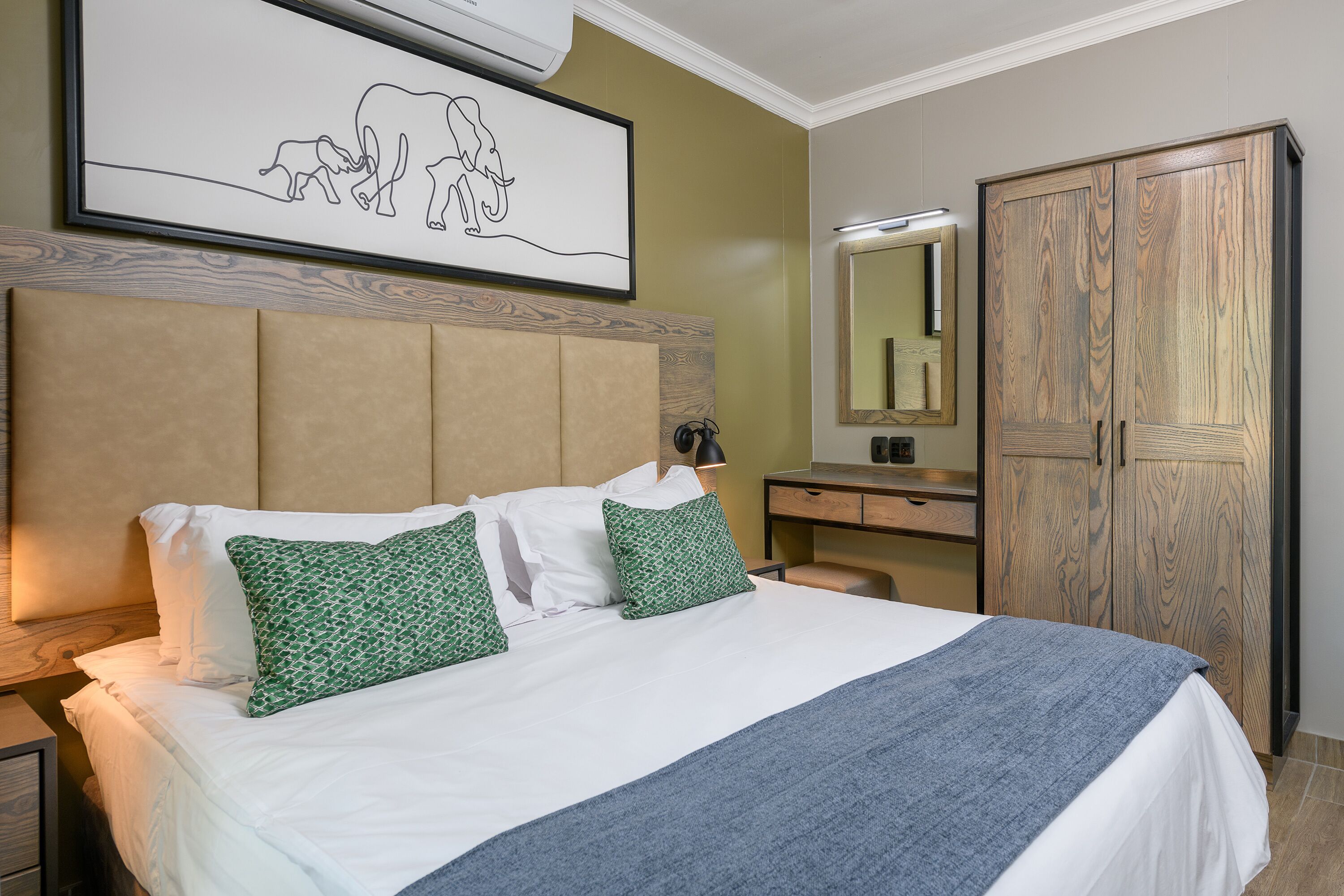 Una cama bien hecha con almohadas blancas y verdes, un cabecero de madera, una mesita de noche con lámpara y un espejo grande sobre un armario de madera.