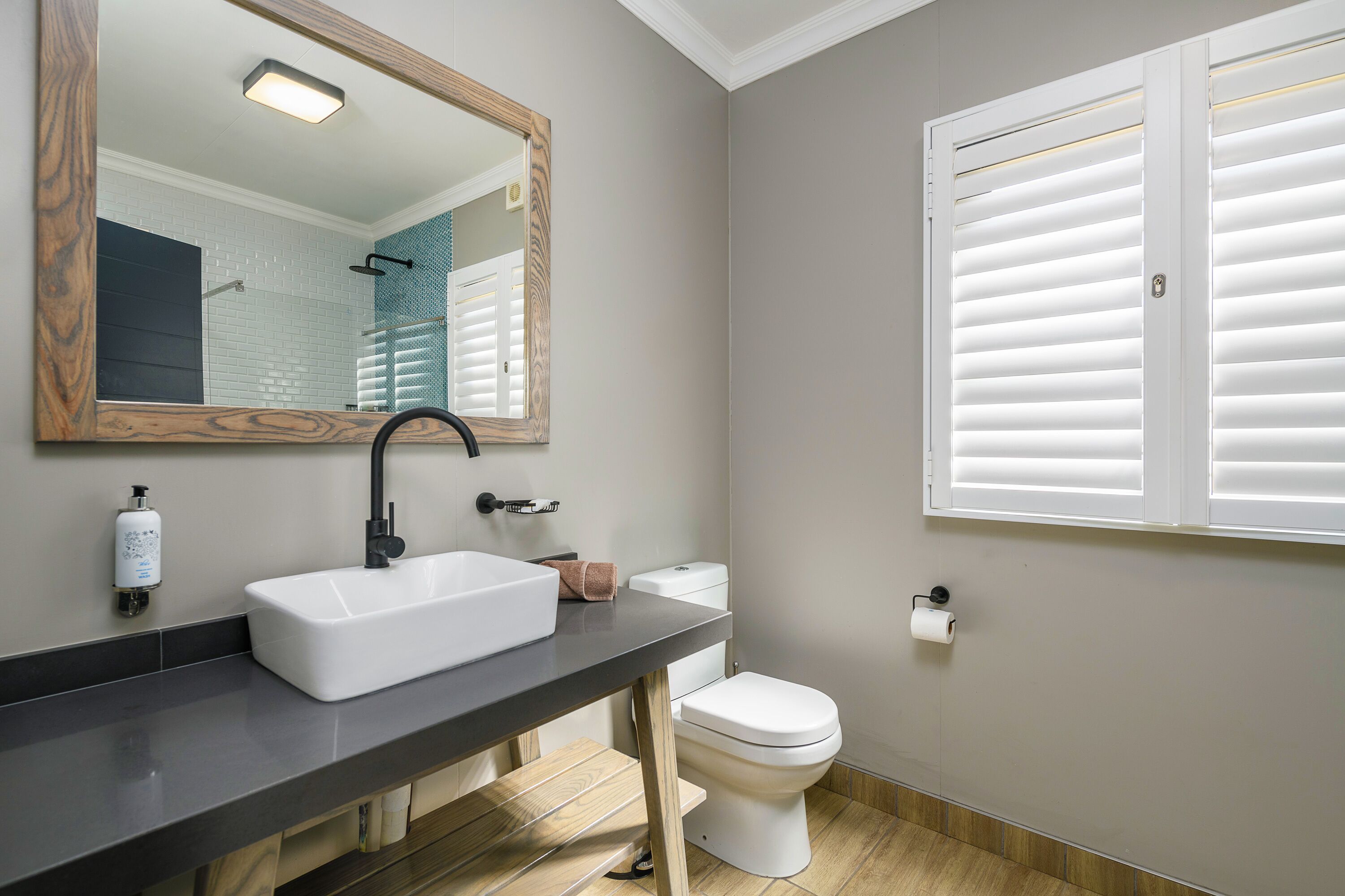 Un bagno con un lavandino bianco, uno specchio con cornice in legno, un water e una finestra con persiane bianche.