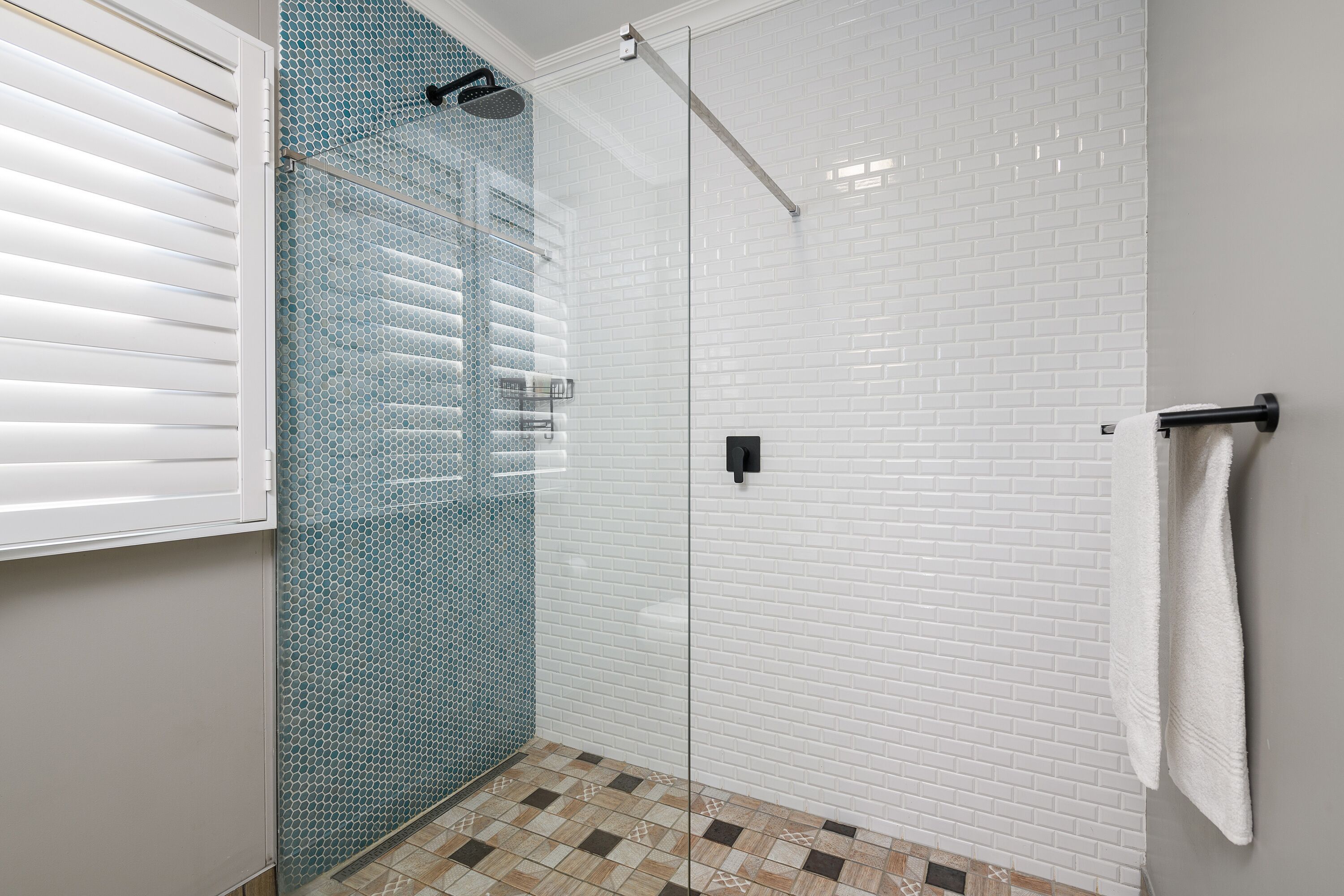 A glass-enclosed shower with white and blue mosaic tiles, a towel rack, and a window with blinds.