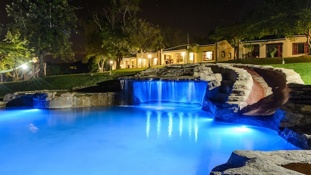 Vue nocturne d’un complexe hôtelier avec une piscine illuminée, une cascade et une série de bâtiments reliés entre eux, dont les fenêtres sont éclairées.