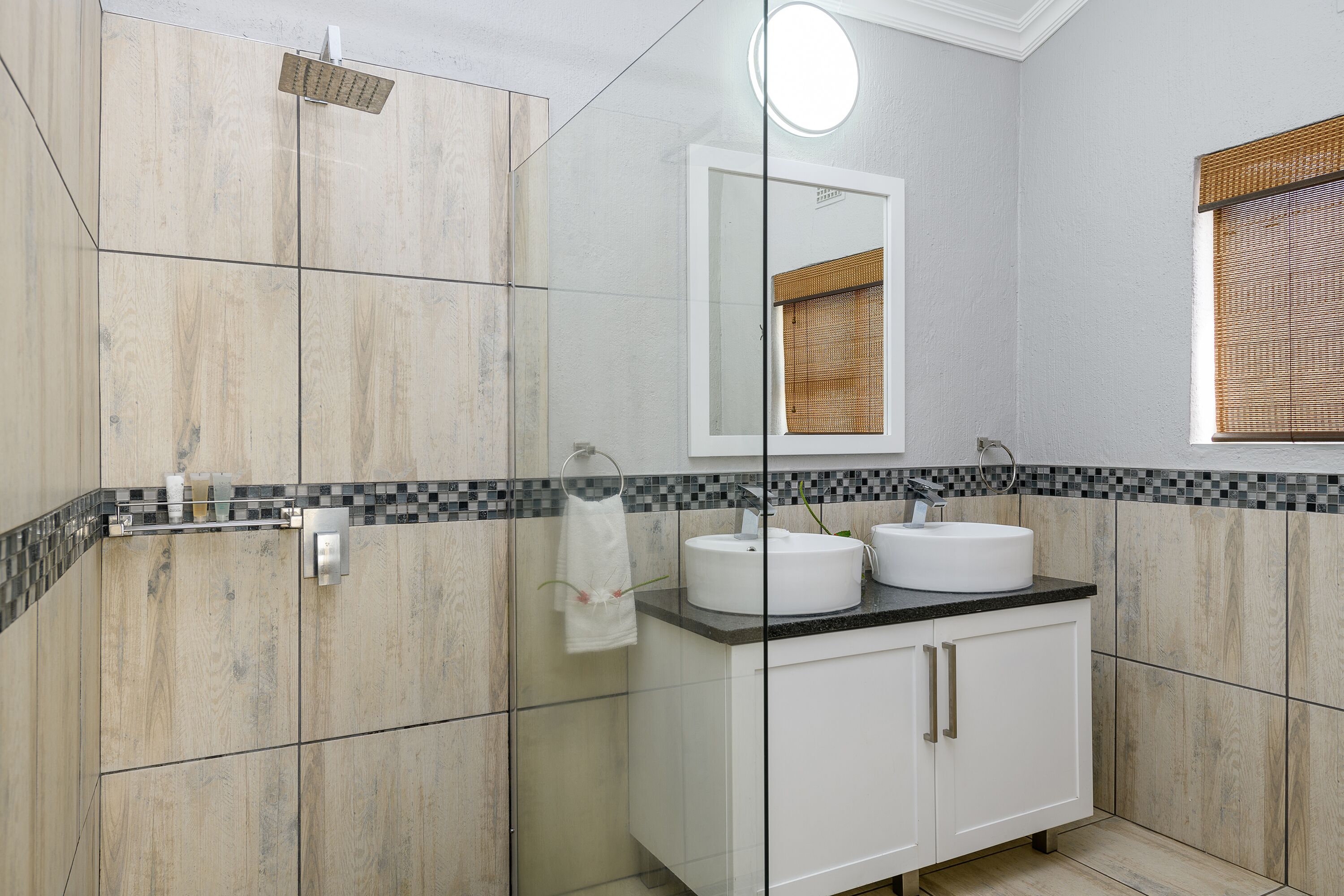 Un baño moderno con cabina de ducha de vidrio, tocador blanco con dos lavamanos y espejo sobre el tocador.
