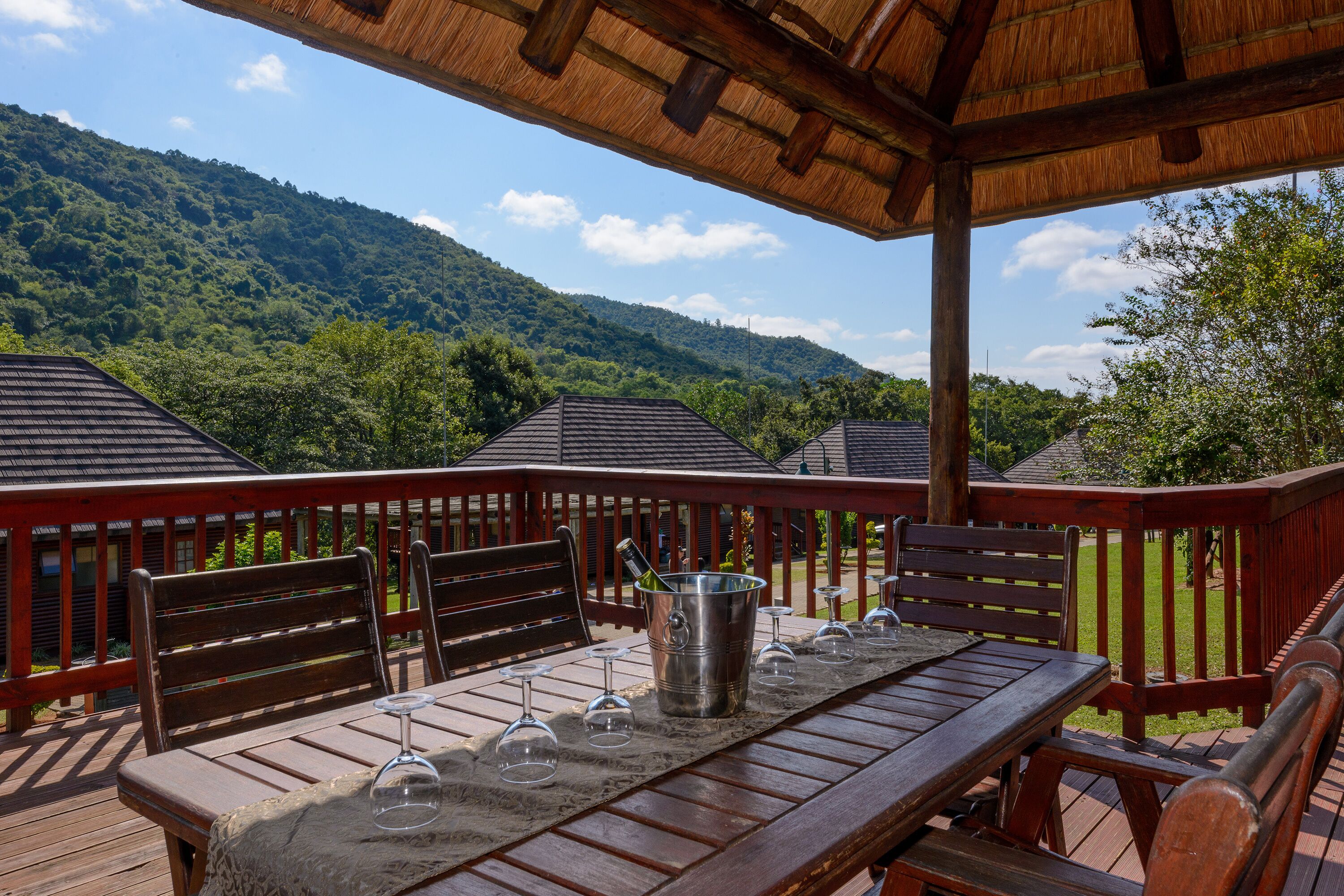 A wooden deck with a table set for a meal, surrounded by chairs, overlooking a lush green landscape with hills in the background.