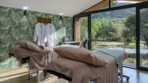 Une salle de spa équipée de deux tables de massage, d’un banc en bois et offrant une vue sur une rivière et une forêt.