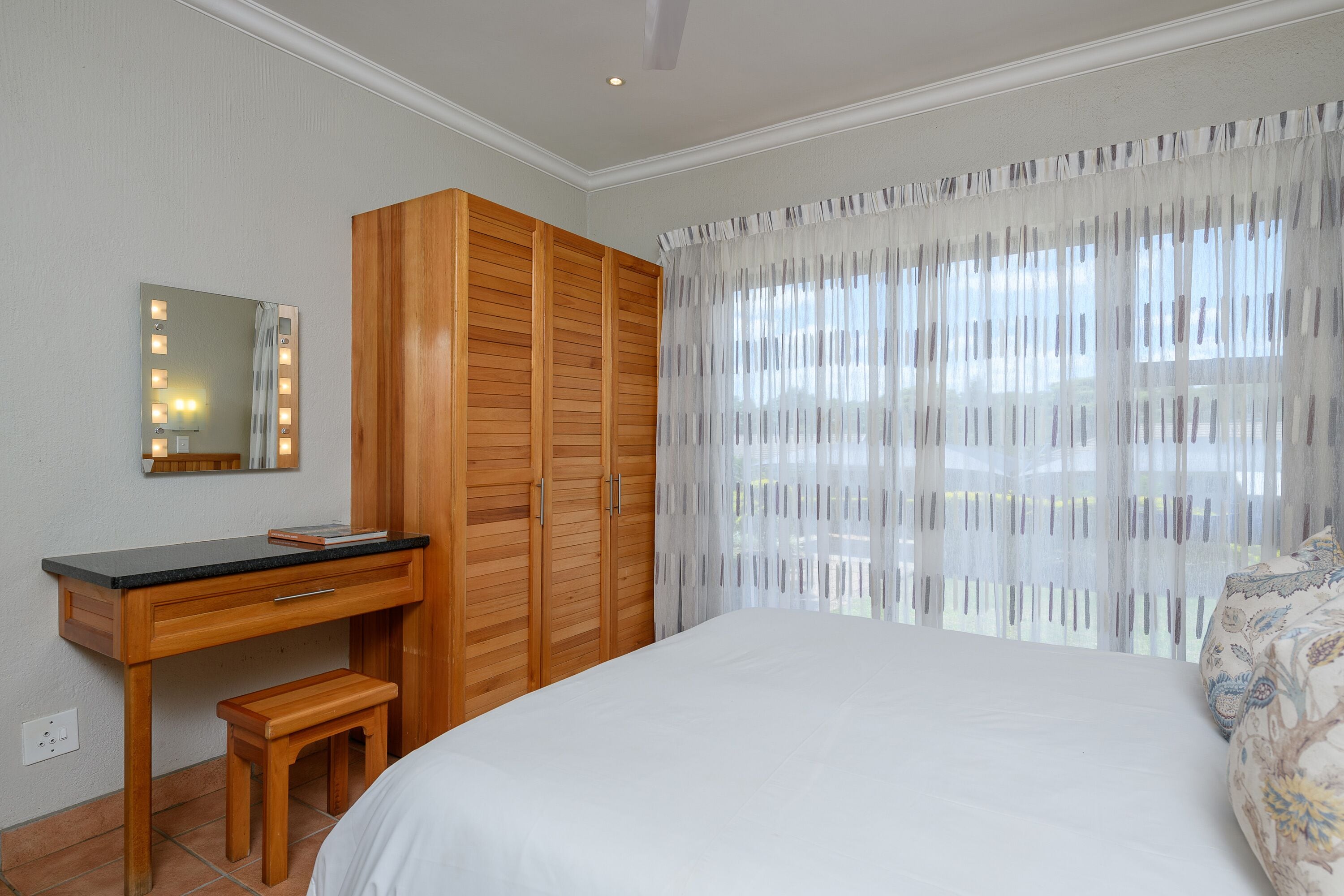 Un dormitorio con un escritorio de madera, un espejo con iluminación, una cama y una ventana con cortinas translúcidas.