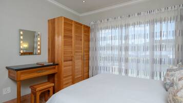 Un dormitorio con un escritorio de madera, un espejo con iluminación, una cama y una ventana con cortinas translúcidas.