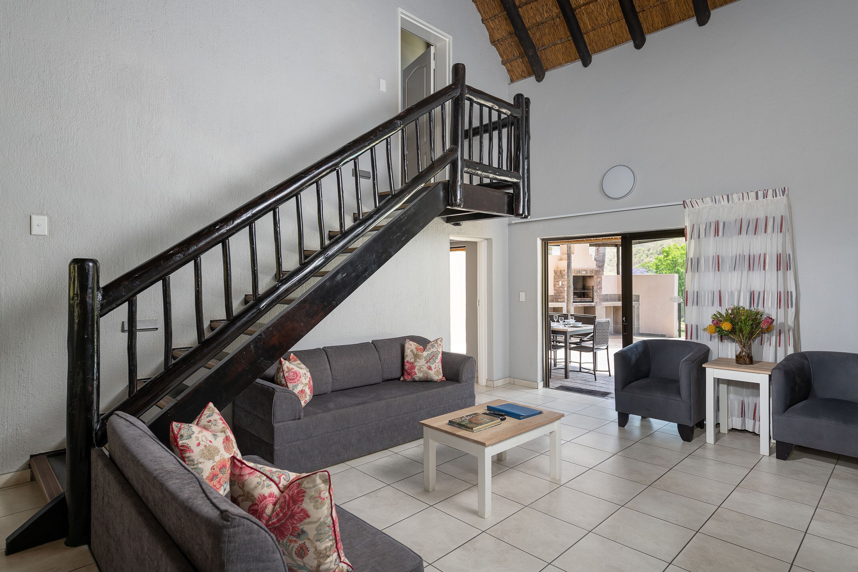 A living room with a staircase, grey sofas, a coffee table, and a dining area visible through an open door.