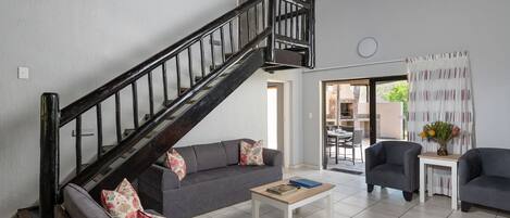 Een woonkamer met een trap, grijze banken, een salontafel en een eetgedeelte zichtbaar door een open deur.