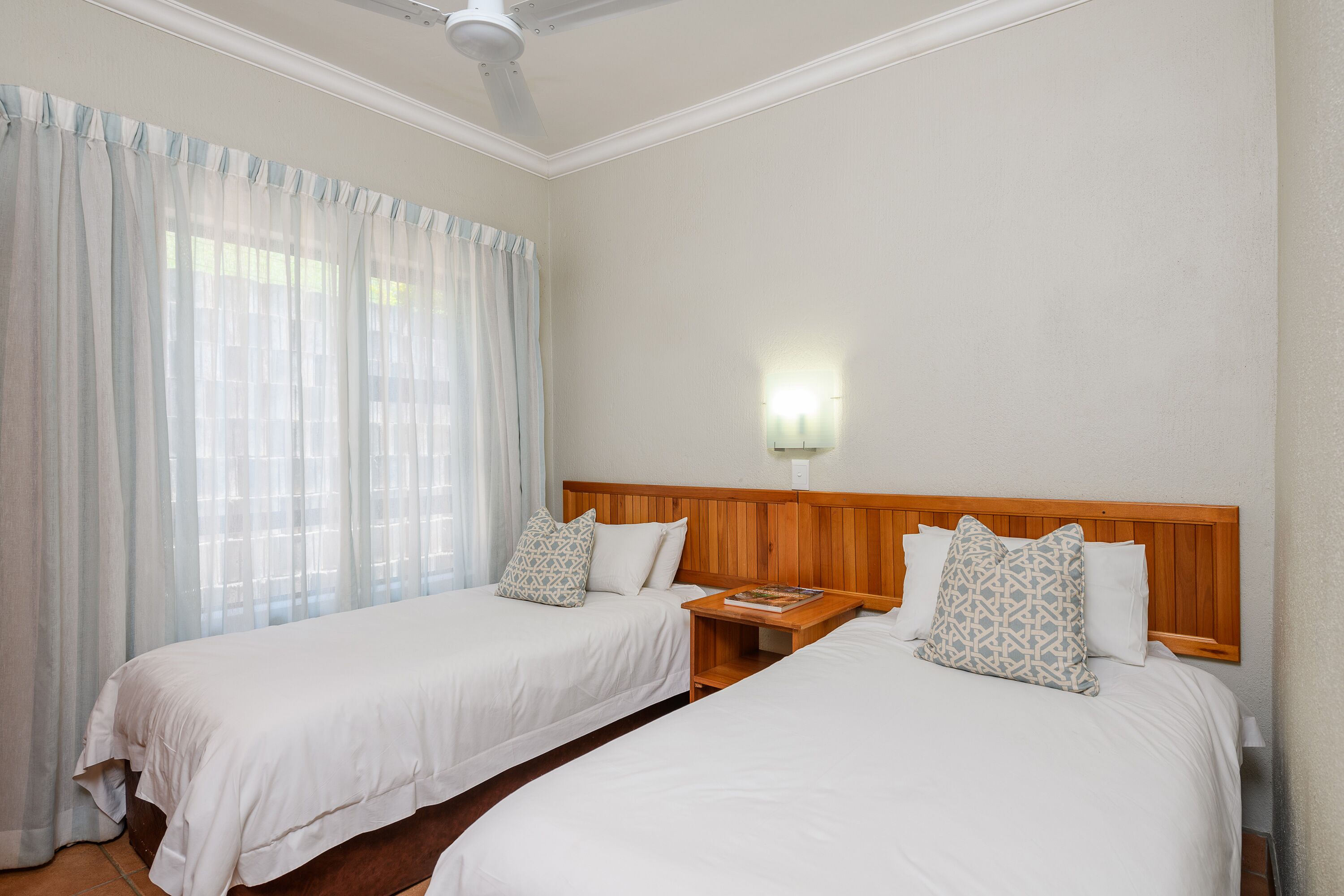A hotel room with two single beds, a wooden headboard, a bedside table with a book, and a wall-mounted light.