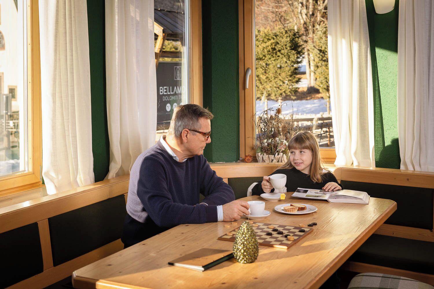 A man and a child at a wooden table with a chessboard, white cups, and a small plant.