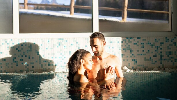 A couple in a swimming pool with a tiled wall and a wooden fence in the background. - Phi Hotel Bellamonte (Predazzo)
