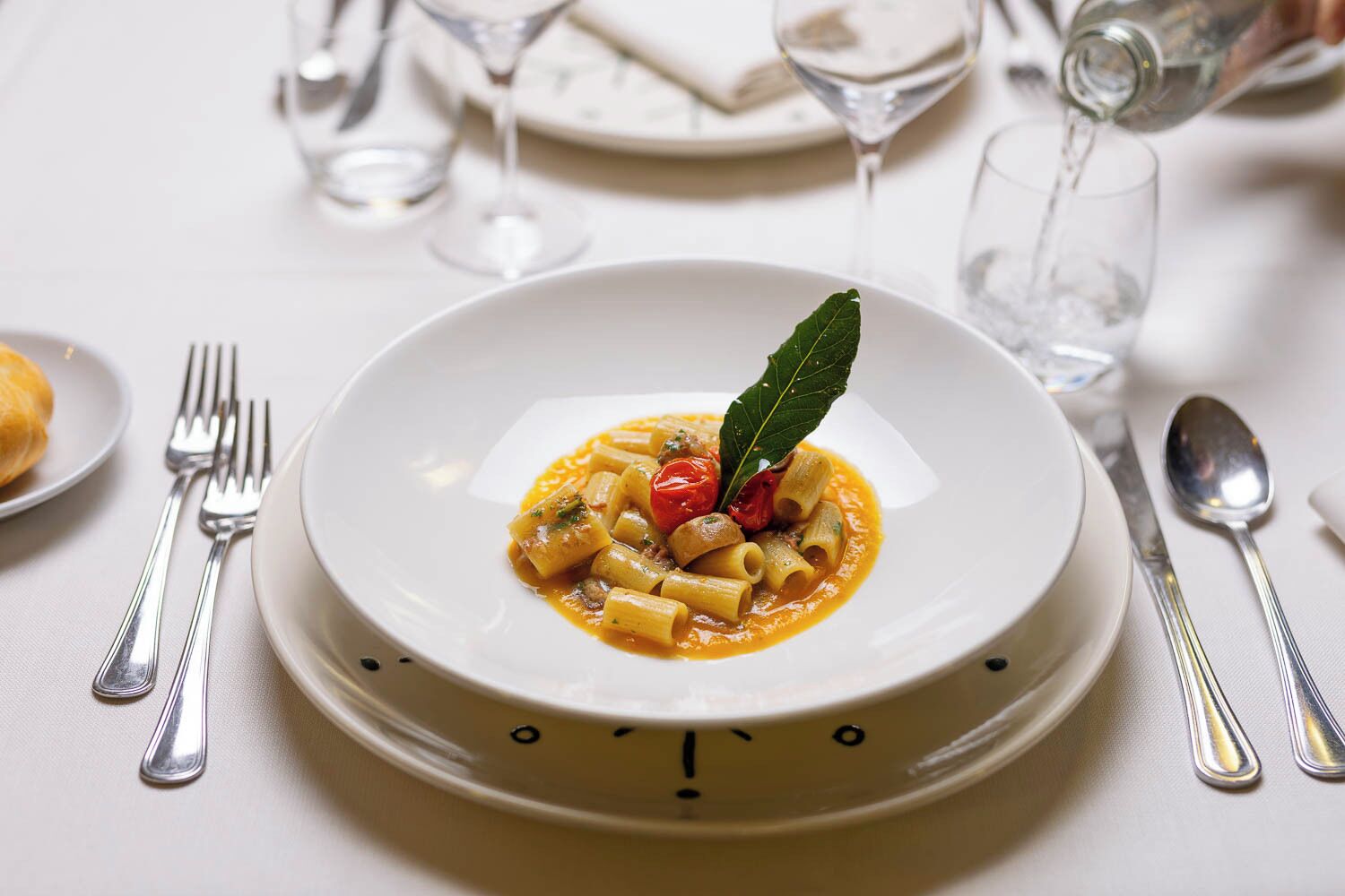 A plate of pasta with a garnish, surrounded by dining utensils and glasses on a table.