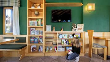 Children can play and learn amidst a vibrant green wall, surrounded by books and toys, while adults relax on wooden benches and tables.