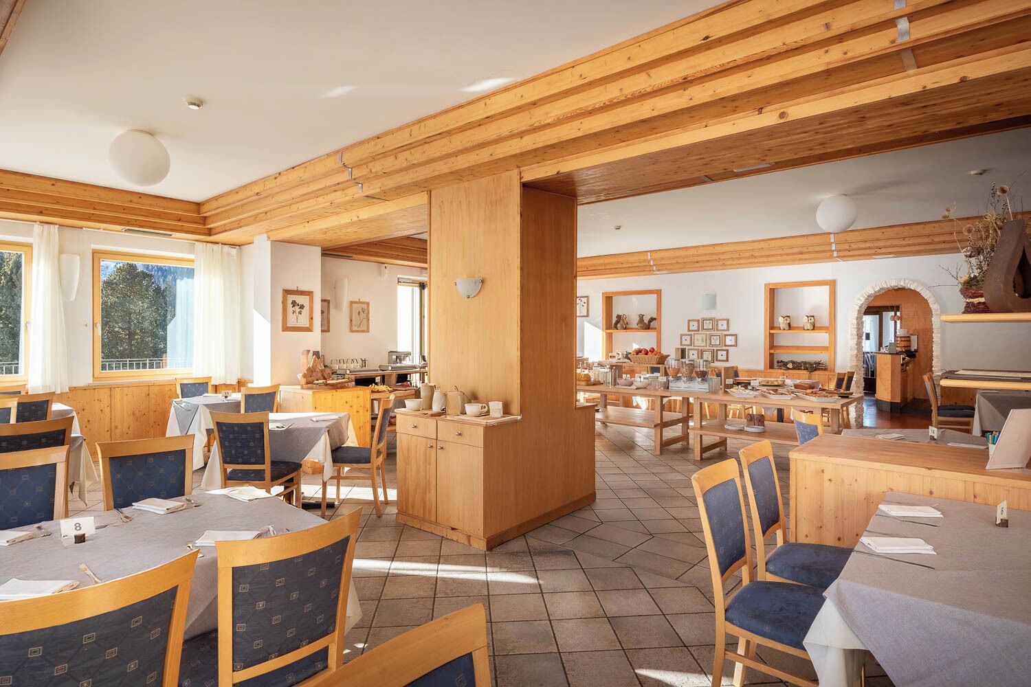 A spacious dining area with wooden tables and chairs, a central pillar, and a kitchen in the background.
