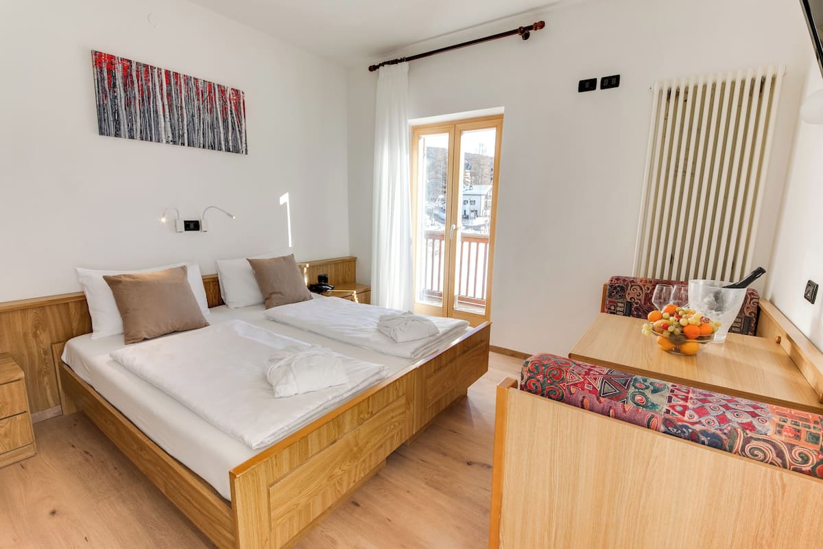 A modern hotel room with a wooden bed, a dining table with a fruit bowl, a colorful bench, and a large window with a view.