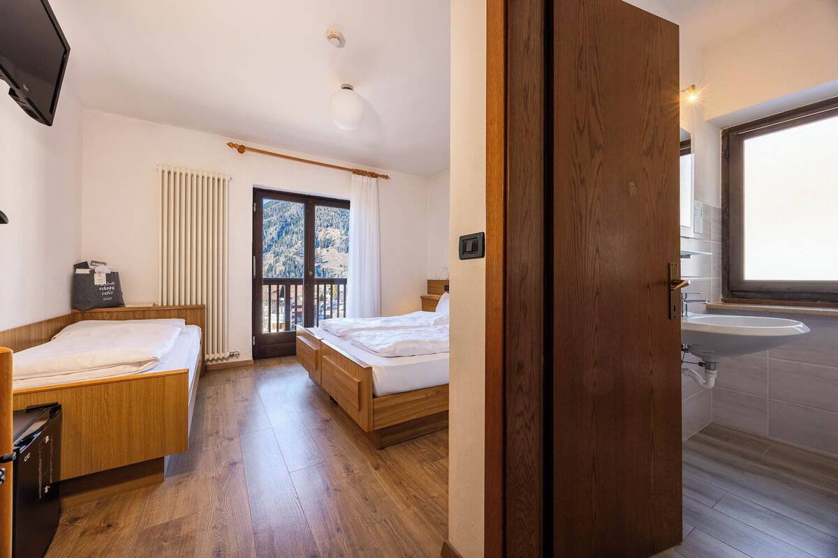 A hotel room with two beds, a wooden floor, a radiator, a window with curtains, and a bathroom with a sink.