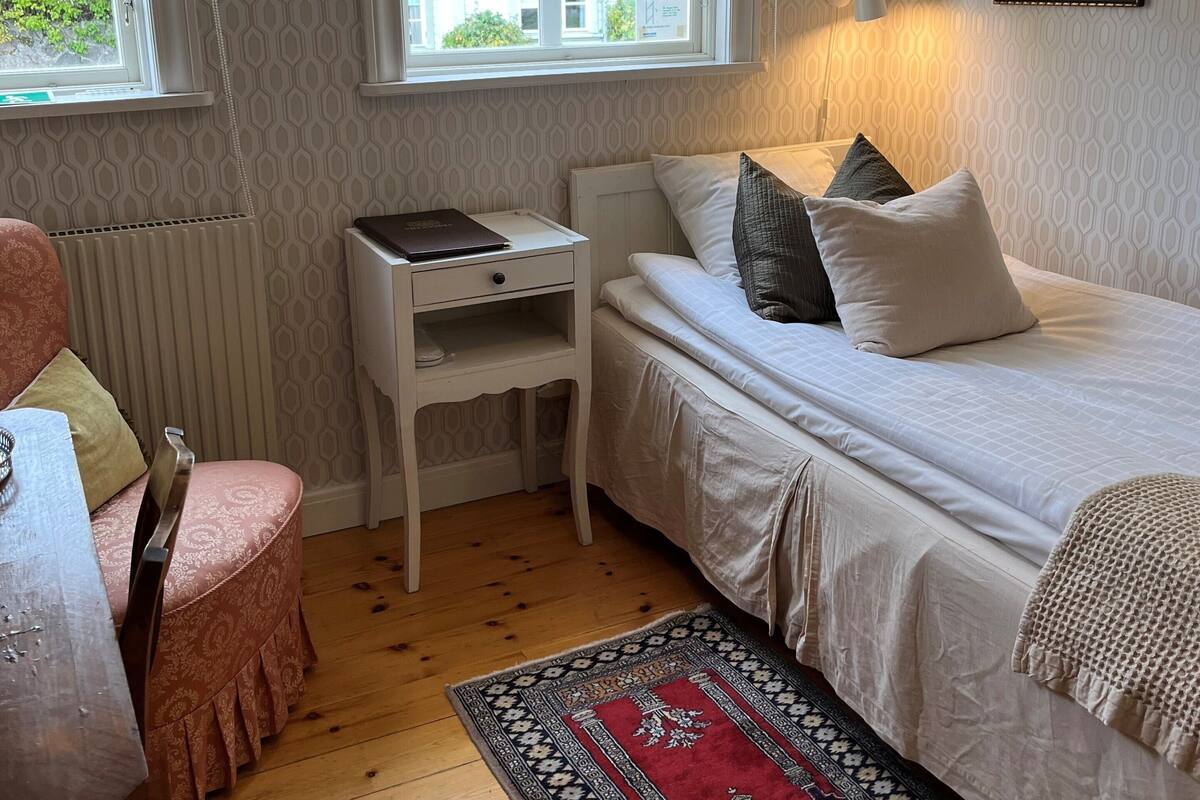 A bedroom with a bed, a desk, a chair, a window with blinds, a radiator, and a patterned rug.