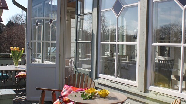 Una terraza de madera con mobiliario exterior, incluyendo sillas y una mesa, y una puerta que da a un espacio cubierto.