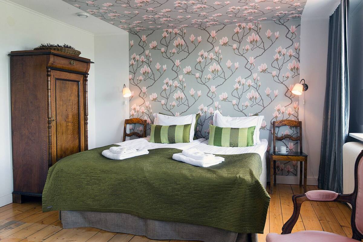 A neatly made bed with green bedding, two bedside tables with lamps, a wooden wardrobe, and floral wallpaper.
