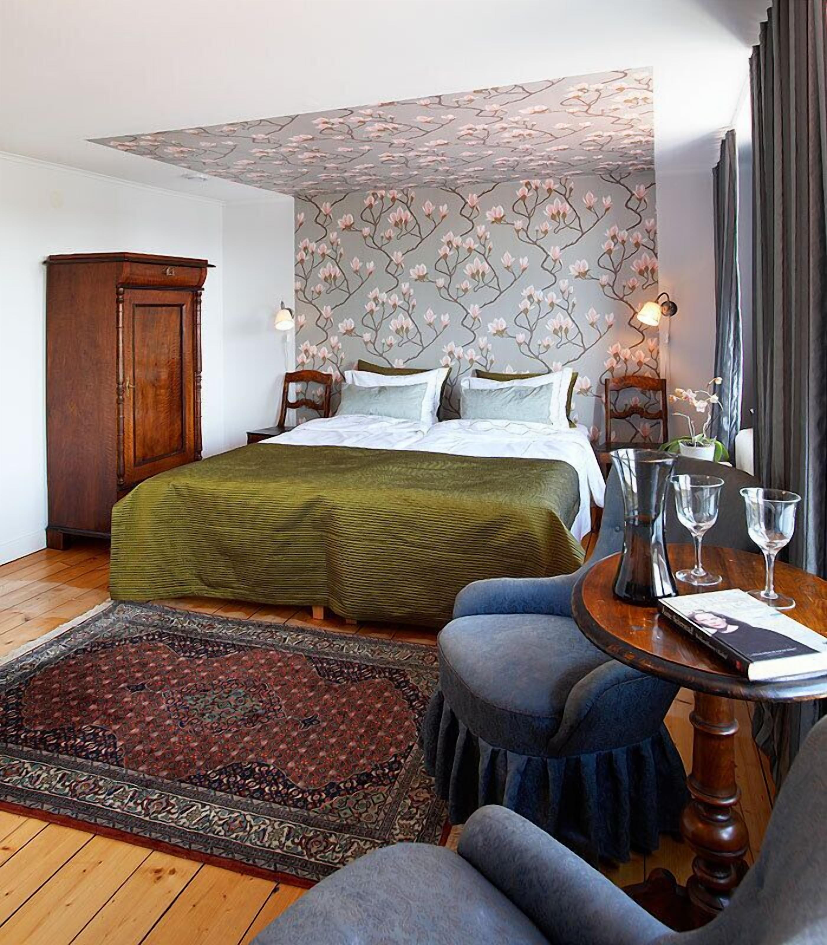 A bedroom with a bed, a wooden wardrobe, a round table with glasses, and a patterned rug.