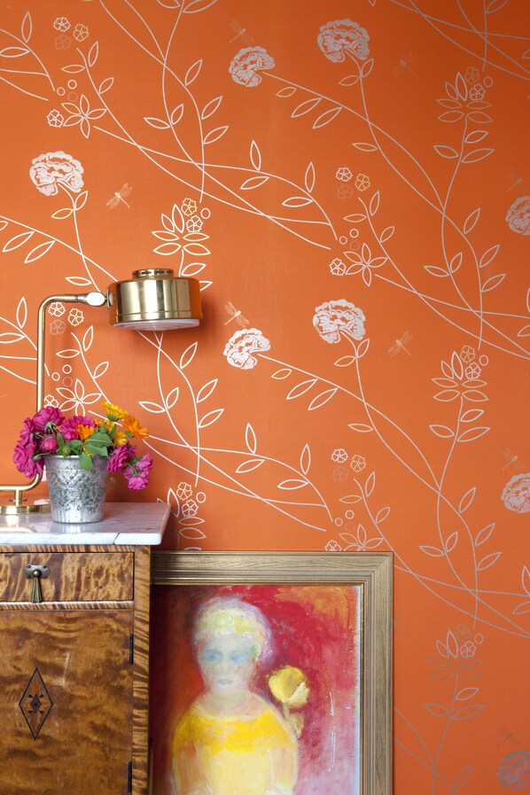 A room with an orange wall featuring white floral patterns, a metal lamp, a vase with flowers, and a framed painting.