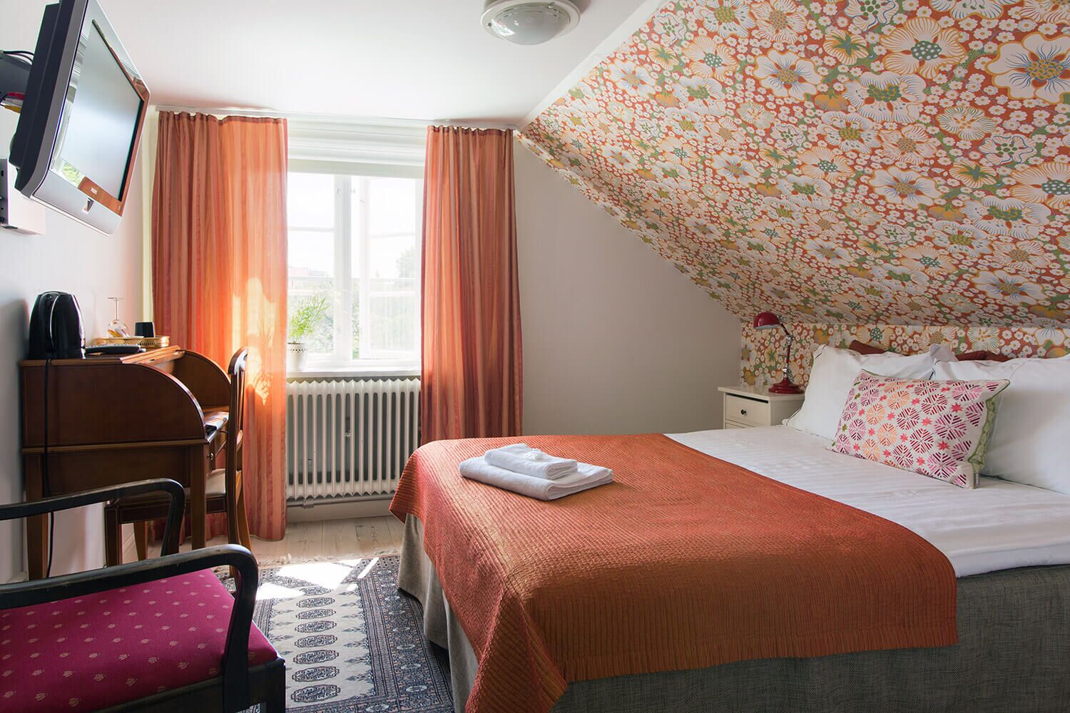 A hotel room with a bed, a desk, a chair, a radiator, and a window with curtains.