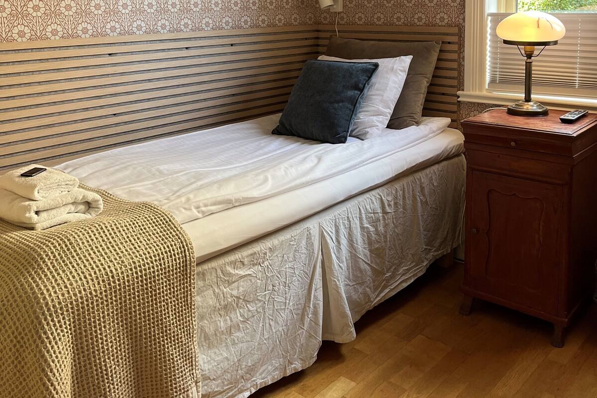 A bedroom with a bed, bedside lamp, wooden nightstand, and patterned wallpaper.
