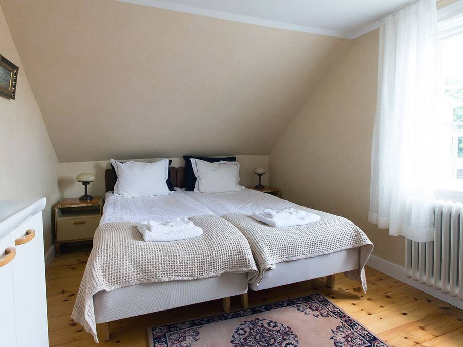 Een tweepersoonsbed met witte lakens, een nachtkastje met een lamp en een raam met gordijnen.