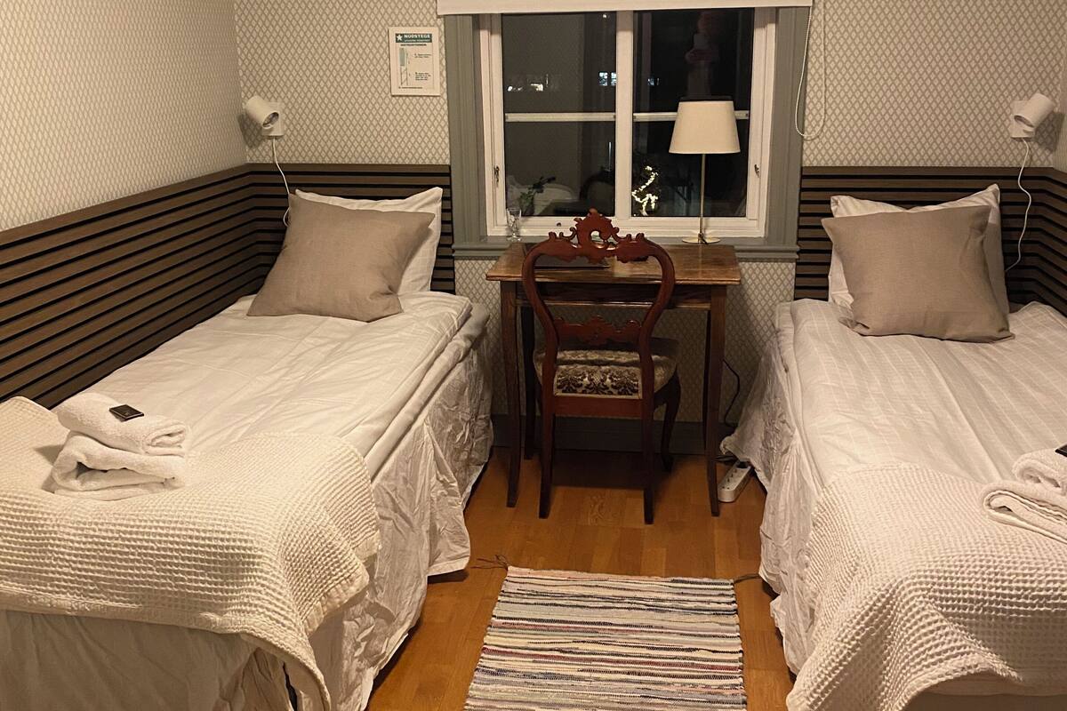 A room with two beds, a wooden desk, a chair, a window, and a floor lamp.