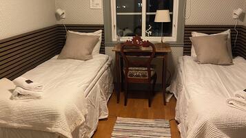 A room with two beds, a wooden desk, a chair, a window, and a floor lamp.