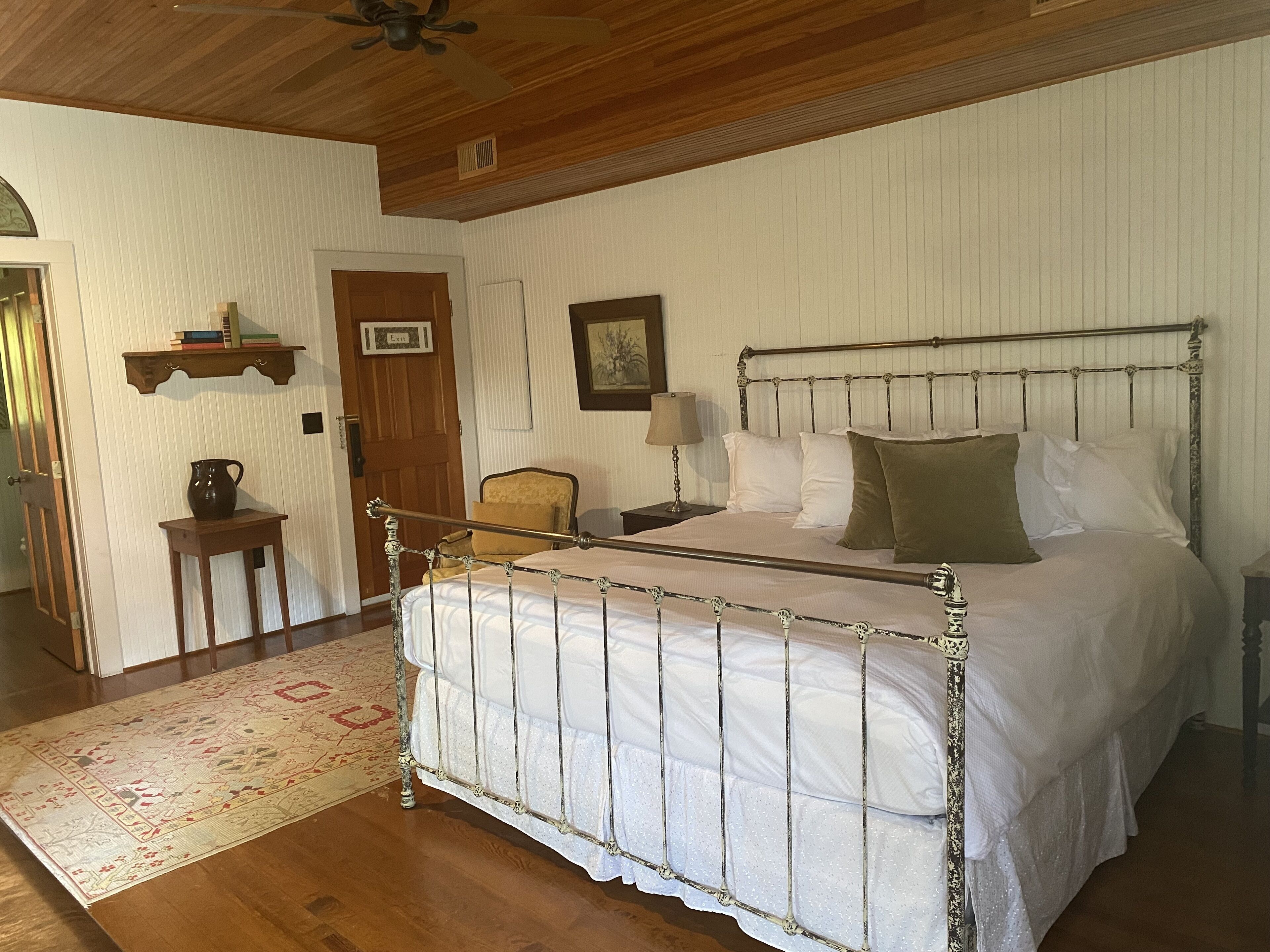 A bedroom with a metal-framed bed, a wooden door, a small table with a pitcher, and a wall-mounted shelf with books.