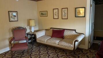 A room with a patterned carpet, a sofa with a red pillow, a chair, a table with a lamp, and framed artwork on the wall.