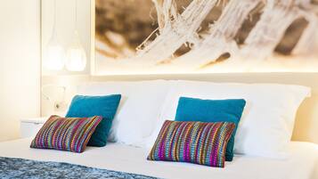 Una cama con sábanas blancas, dos almohadas turquesa y un colorido mantón tejido.