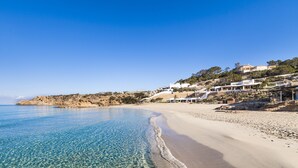A beach with clear blue water, sandy shore, and buildings on a hillside. - Insotel Tarida Beach Resort & SPA - All inclusive (Sant Josep de sa Talaia)