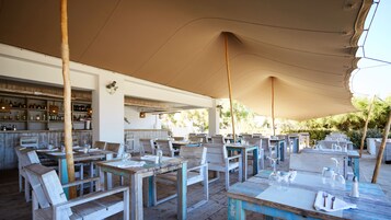 An outdoor dining area with wooden tables and chairs, a bar with shelves of bottles, and a covered section with a beige canopy.