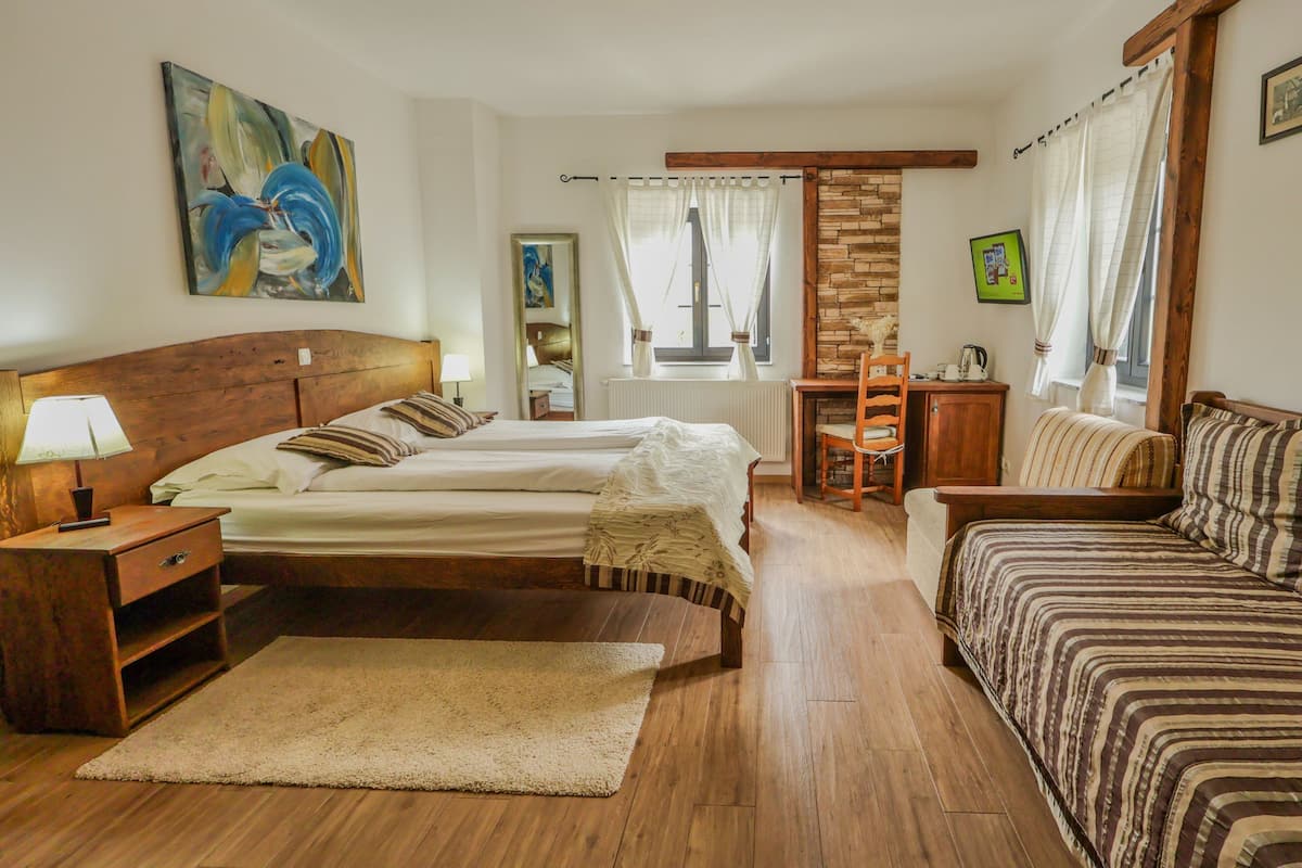 A room with a bed, a sofa, a wooden nightstand, a lamp, a radiator, a window with curtains, a painting, and a brick wall.