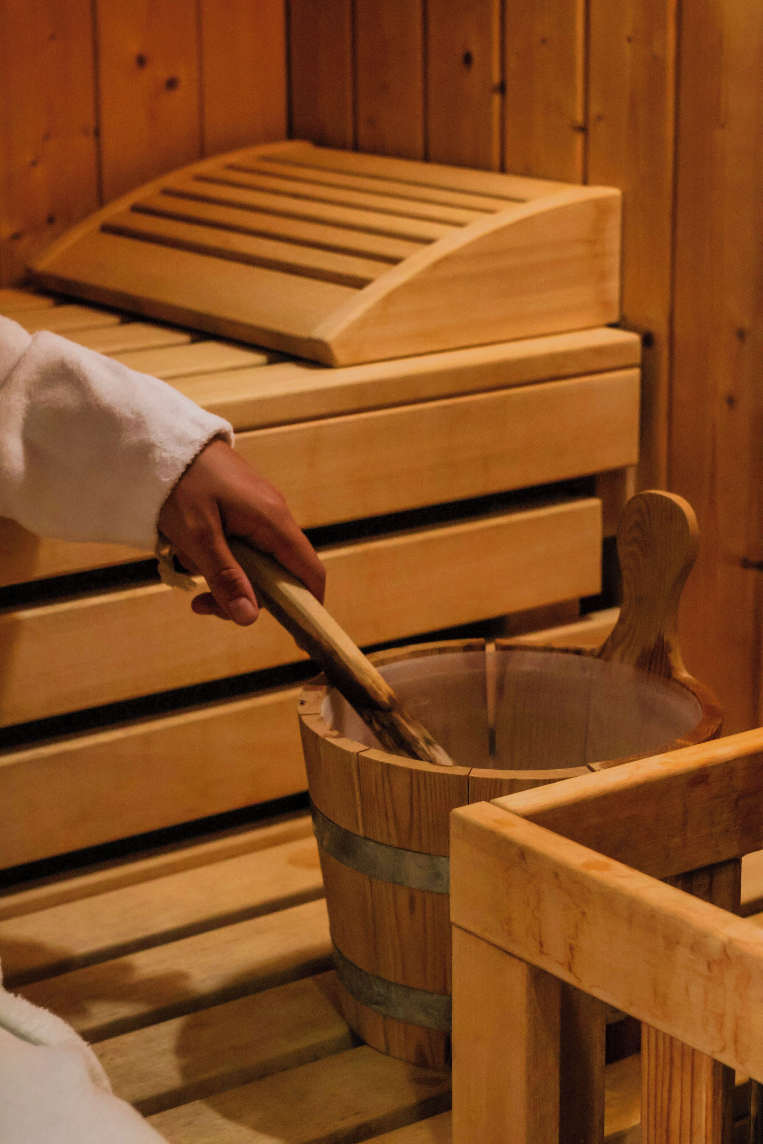 Un banc en bois dans un sauna, sur lequel est assis un individu qui utilise une louche en bois.
