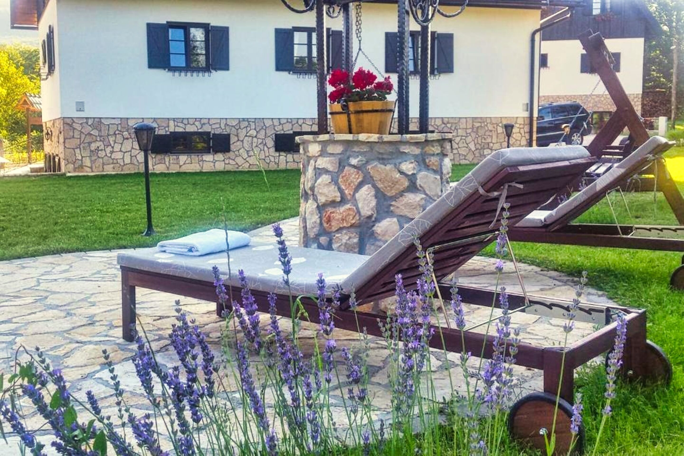 Een terras van natuursteen met twee loungestoelen, een stenen tafel en een hangplanter met rode bloemen.