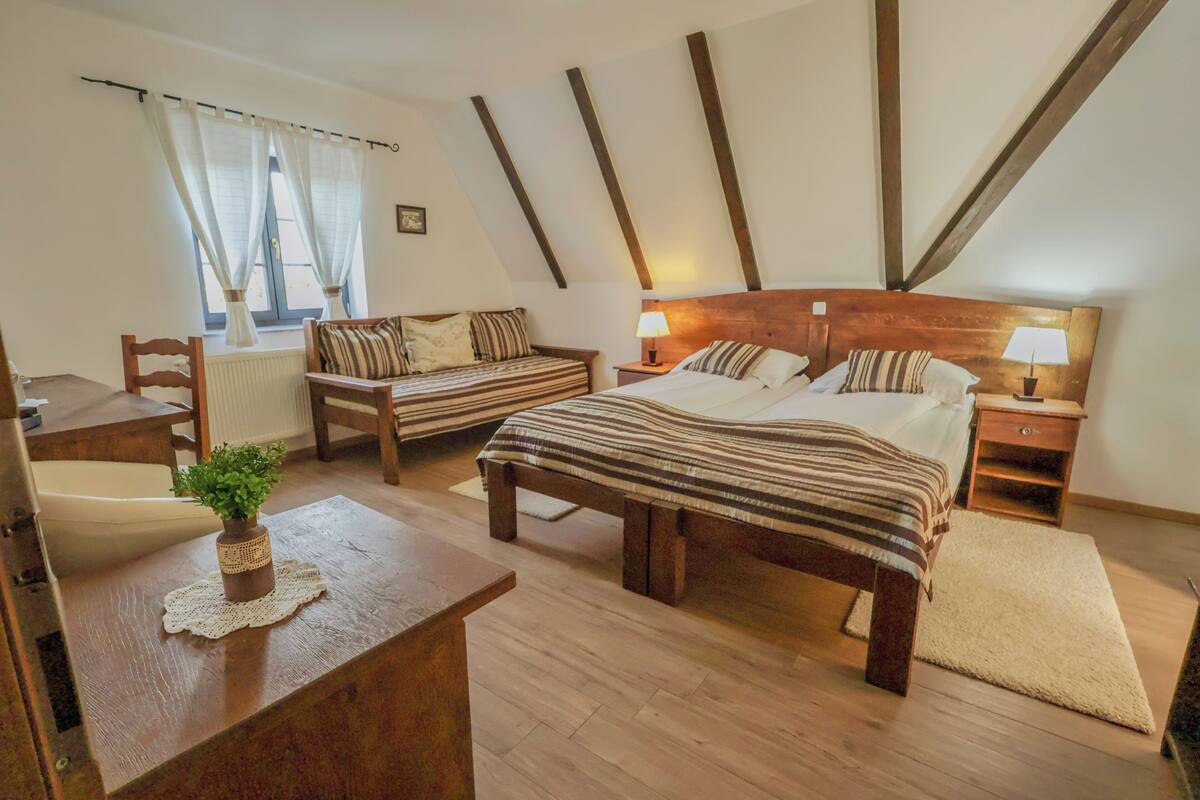 A bedroom with a wooden bed, a wooden nightstand, a bedside table, a chair, a small table with a plant, and a window with curtains.