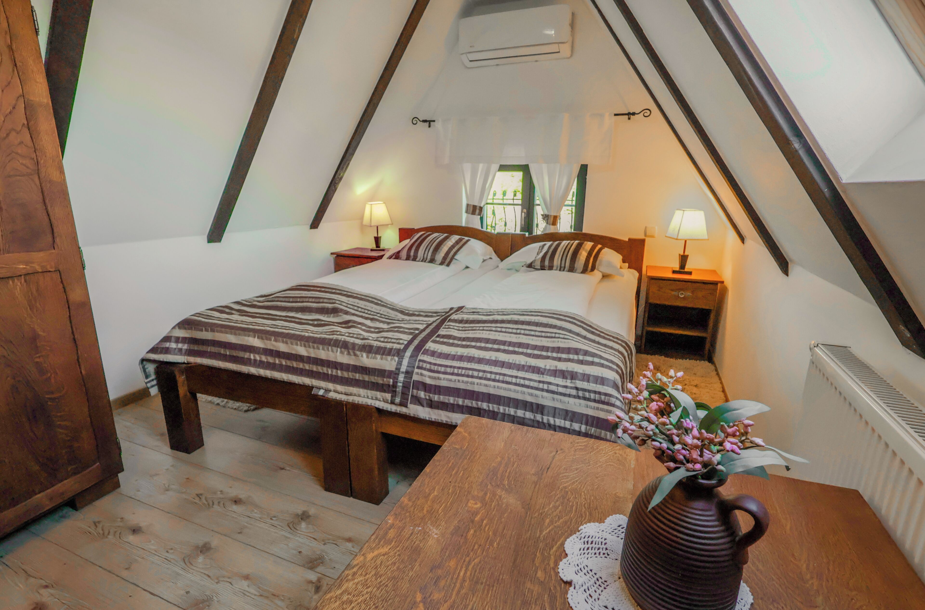 Una camera da letto con soffitto spiovente, un letto di legno, lampade sui comodini, un tavolino e un vaso con fiori.