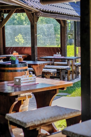 An outdoor dining area with wooden tables and benches, a covered roof, and a view of greenery.