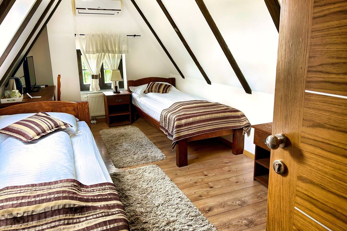 A bedroom with two beds, a wooden floor, a window with curtains, a desk with a lamp, and an air conditioning unit.