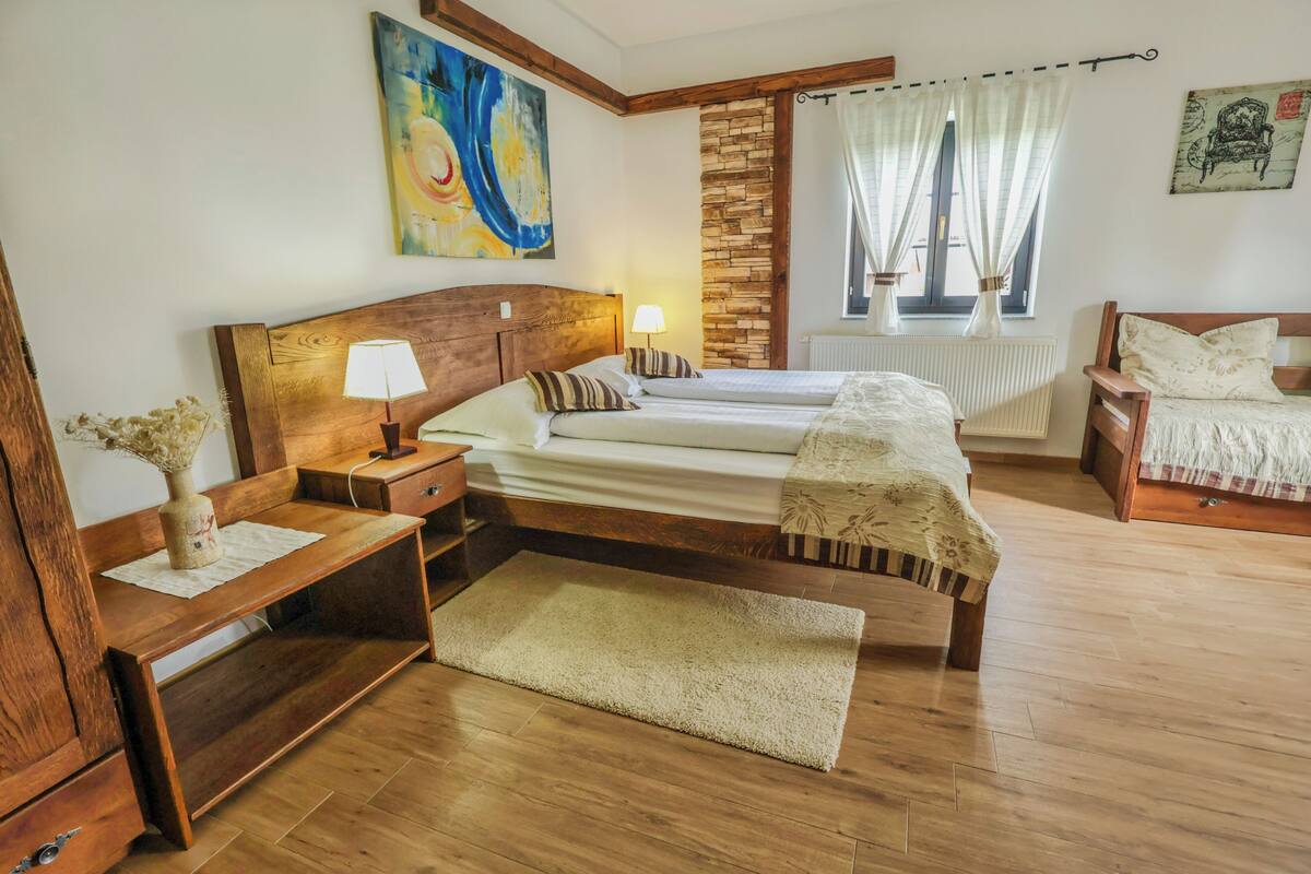 A bedroom with a wooden headboard, two beds, a nightstand, a bedside table, a lamp, a vase with dried flowers, a window with curtains, and a radiator.