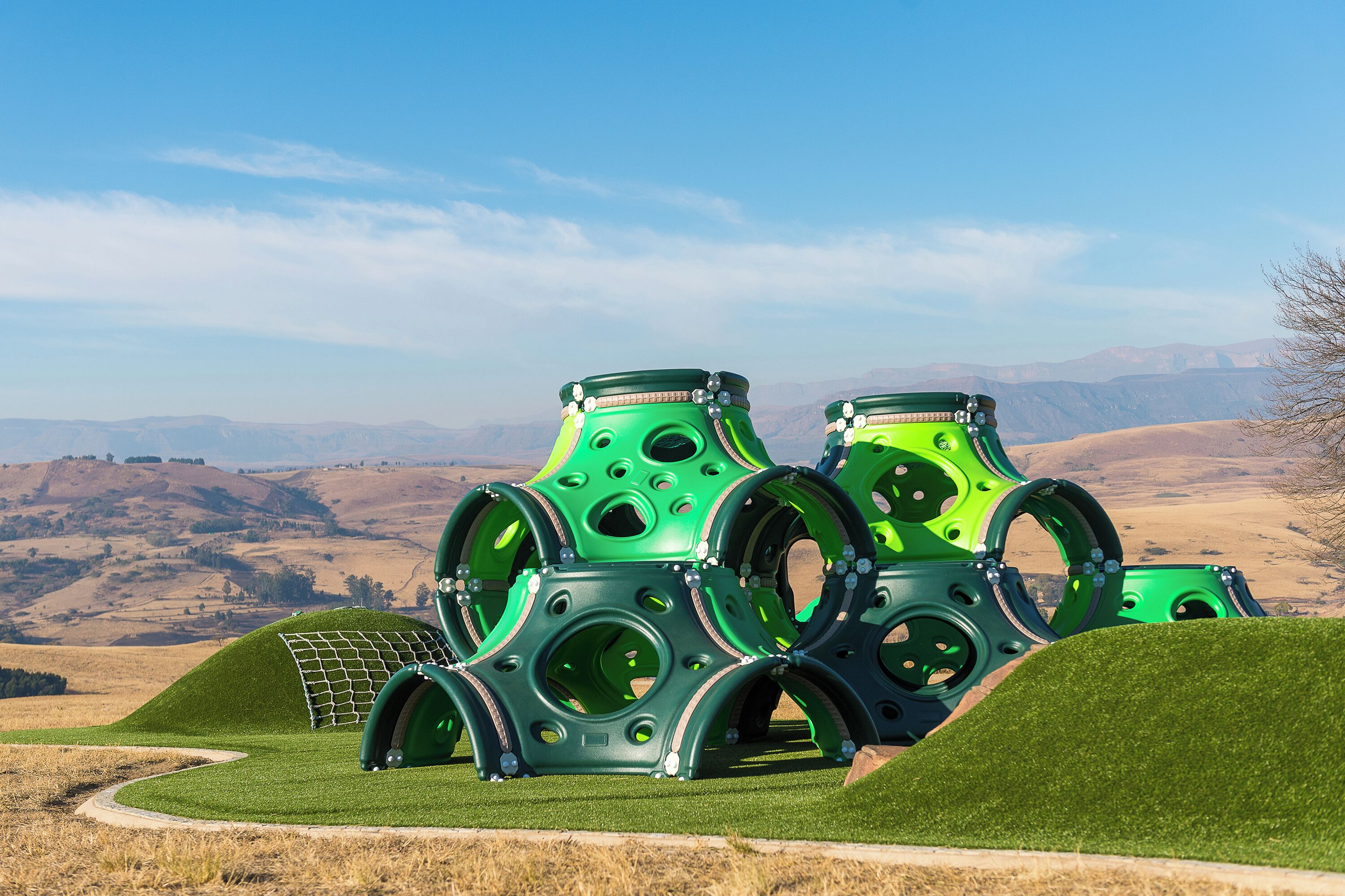 A green, spherical structure with multiple openings, surrounded by a grassy area with a hilly landscape in the background.