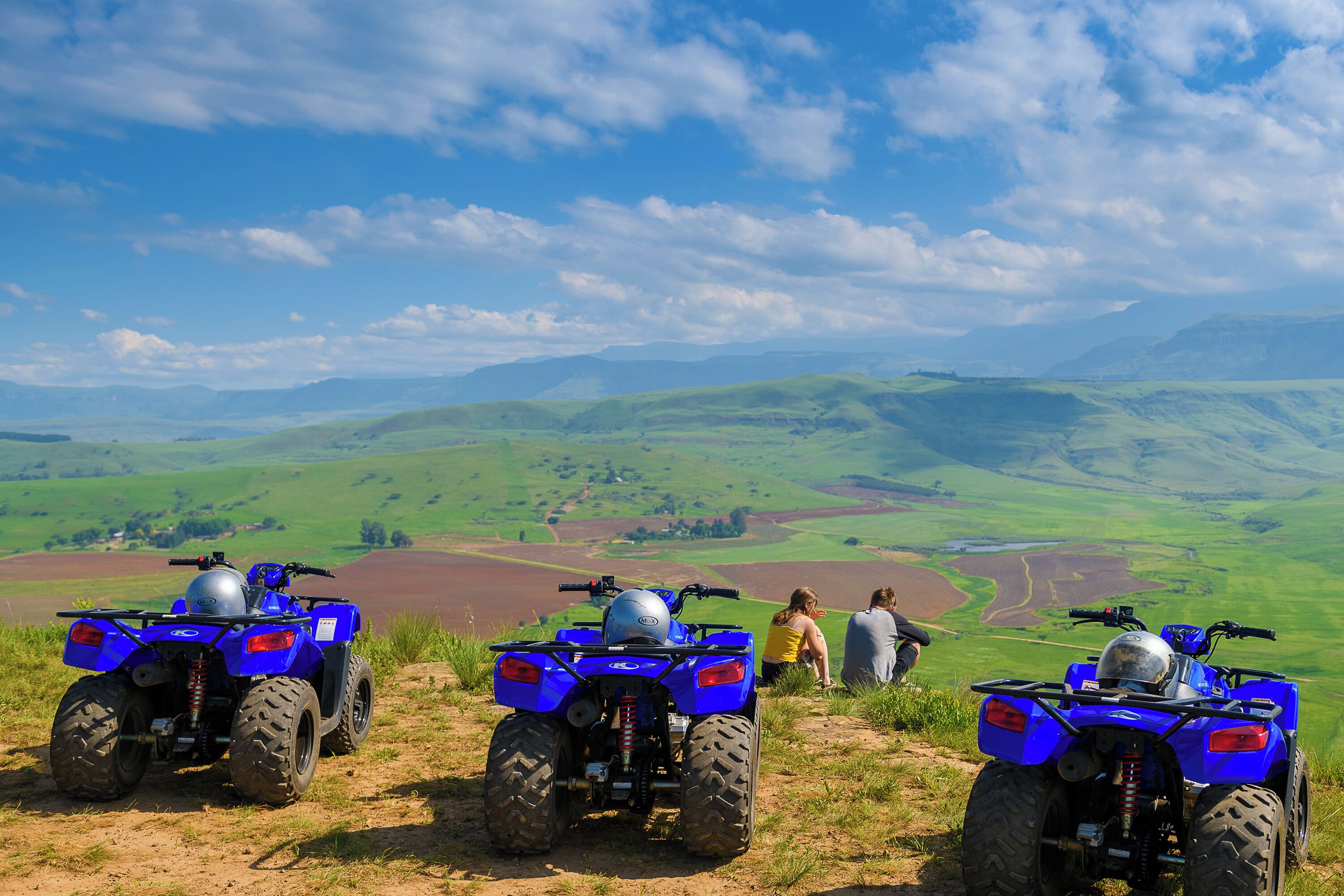 Drei blaue Quads auf einem grasbewachsen Hügel mit Blick auf grüne Felder und Hügel im Hintergrund.